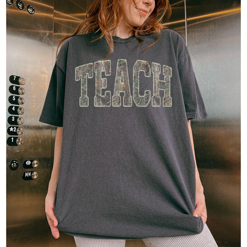 May include: Dark gray t-shirt with the word "TEACH" in large, textured letters across the front. The letters have a camouflage-like pattern. The shirt has short sleeves and a relaxed fit.