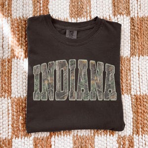 K&ouml;nnte beinhalten: Dunkelbraunes T-Shirt mit dem Wort "INDIANA" in gro&szlig;en Blockbuchstaben auf der Vorderseite. Die Buchstaben sind mit einem Camouflagemuster gef&uuml;llt und wei&szlig; umrandet. Das Shirt ist ordentlich auf einer strukturierten, neutral get&ouml;nten Oberfl&auml;che gefaltet.