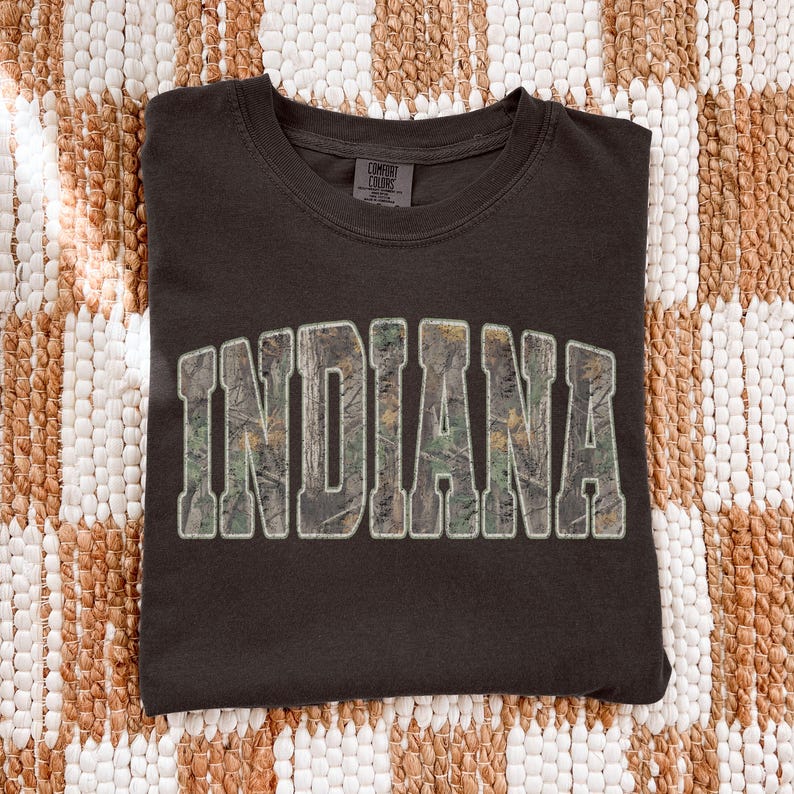 K&ouml;nnte beinhalten: Dunkelbraunes T-Shirt mit dem Wort "INDIANA" in gro&szlig;en Blockbuchstaben auf der Vorderseite. Die Buchstaben sind mit einem Camouflagemuster gef&uuml;llt und wei&szlig; umrandet. Das Shirt ist ordentlich auf einer strukturierten, neutral get&ouml;nten Oberfl&auml;che gefaltet.