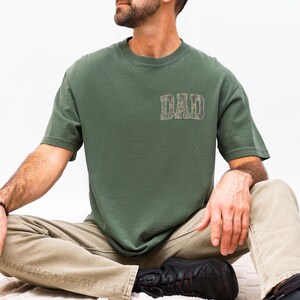 May include: A short-sleeved olive green t-shirt featuring the word DAD printed in a camouflage pattern on the upper left chest. The shirt has a crew neckline and a standard fit, constructed from a solid-colored fabric with a simple design.