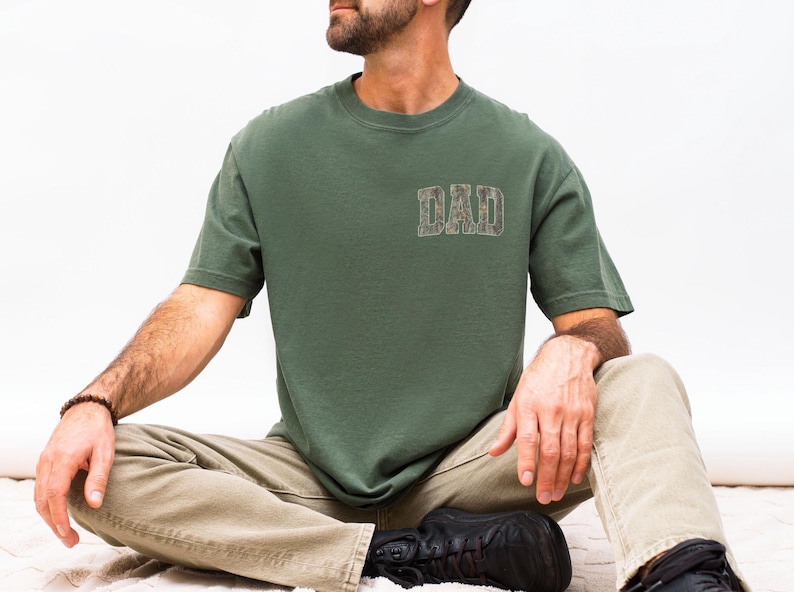 May include: A short-sleeved olive green t-shirt featuring the word DAD printed in a camouflage pattern on the upper left chest. The shirt has a crew neckline and a standard fit, constructed from a solid-colored fabric with a simple design.