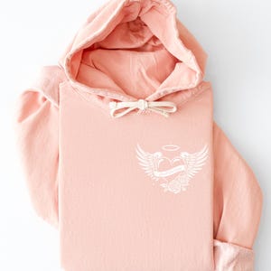 May include: A folded, light pink hoodie with a drawstring hood and a white graphic on the chest. The graphic features a heart with wings, a halo, and the text "Love Over Doubt". The sleeves are cuffed.