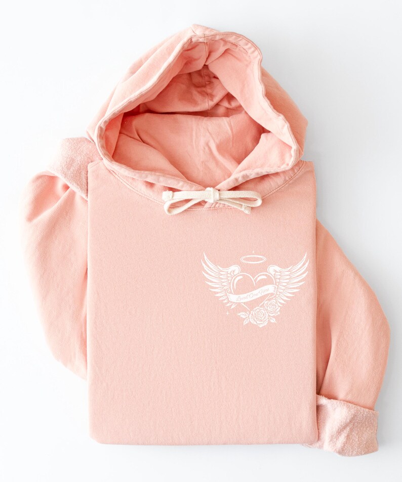 May include: A folded, light pink hoodie with a drawstring hood and a white graphic on the chest. The graphic features a heart with wings, a halo, and the text "Love Over Doubt". The sleeves are cuffed.