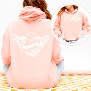 May include: A light pink hoodie with a white graphic on the back featuring a heart with angel wings, a halo, and roses. The text "Loved One Here" is on a banner across the heart. A smaller version of the graphic is on the front.