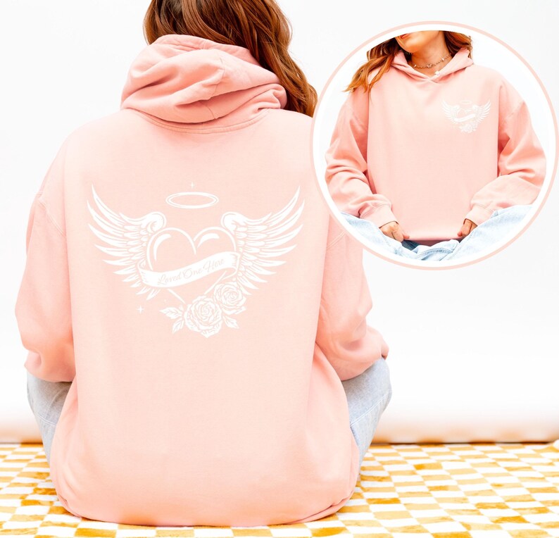 May include: A light pink hoodie with a white graphic on the back featuring a heart with angel wings, a halo, and roses. The text "Loved One Here" is on a banner across the heart. A smaller version of the graphic is on the front.