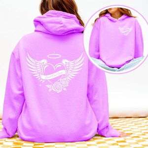 May include: A light purple hoodie with a white graphic on the back featuring a heart, wings, halo, and roses. The text "Loved One Here" is on a banner across the heart. A smaller version of the graphic is on the front.