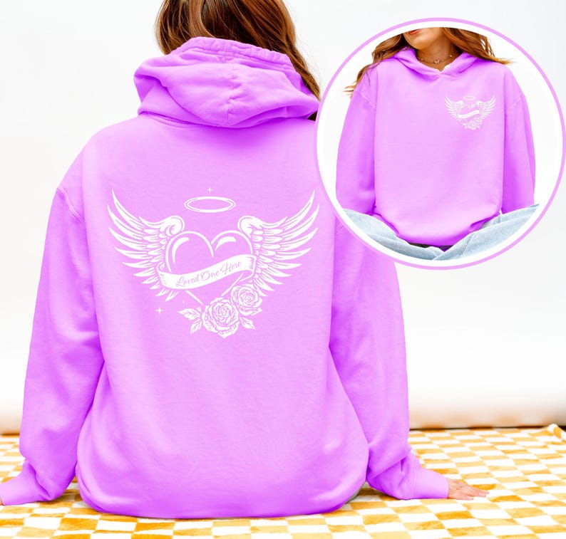 May include: A light purple hoodie with a white graphic on the back featuring a heart, wings, halo, and roses. The text "Loved One Here" is on a banner across the heart. A smaller version of the graphic is on the front.