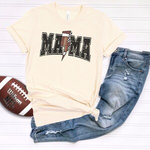 May include: A beige t-shirt with a black and white checkered football graphic and the text "MAMA" in a black distressed font.