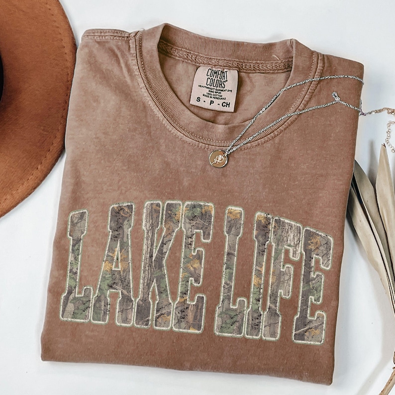 K&ouml;nnte beinhalten: Ein braunes Comfort Colors T-Shirt mit dem Schriftzug "LAKE LIFE" in einem Camouflage-Design. Eine silberne Halskette mit einem kleinen Anh&auml;nger liegt &uuml;ber dem Shirt. Ein brauner Hut ist in der oberen linken Ecke zu sehen.