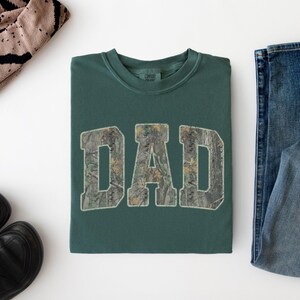 May include: A dark green crewneck t-shirt featuring the word DAD printed in a camouflage pattern across the chest. The letters are in a bold, block-style font with a light-colored outline, centered on the front of the folded garment.