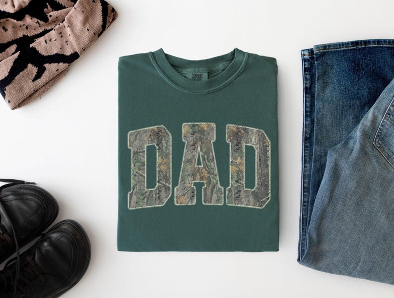 May include: A dark green crewneck t-shirt featuring the word DAD printed in a camouflage pattern across the chest. The letters are in a bold, block-style font with a light-colored outline, centered on the front of the folded garment.