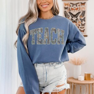 May include: A blue long-sleeve sweatshirt with the word "TEACH" in a camouflage pattern across the chest. The sweatshirt is paired with denim shorts. The overall aesthetic is casual and comfortable.