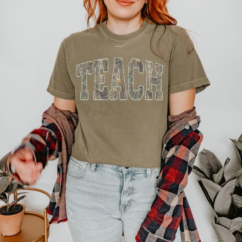 May include: Olive green t-shirt with the word "TEACH" in a camouflage pattern. The shirt is paired with light wash jeans and a red and black plaid flannel shirt. A necklace is visible.