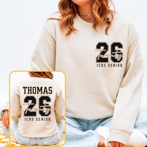May include: A cream-colored sweatshirt with the number "26" in black and the word "twenty" in cursive. The text "ICHS SENIOR" is below the number. A smaller image shows the back of the sweatshirt with the name "THOMAS" above the number.
