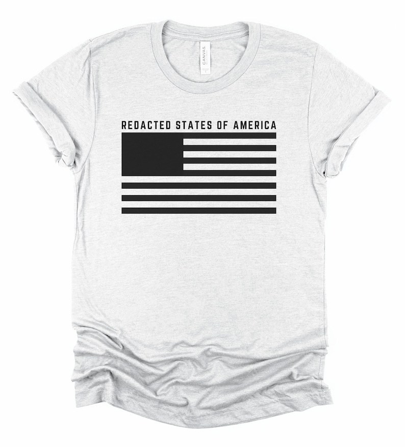 K&ouml;nnte beinhalten: Hellgraues T-Shirt mit dem Text "REDACTED STATES OF AMERICA" &uuml;ber einem schwarz-wei&szlig;en Flaggen-Design. Das Shirt besteht aus einem weichen Material und hat einen Rundhalsausschnitt.