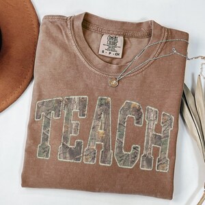 May include: A brown Comfort Colors t-shirt with the word "TEACH" in large, camouflage-patterned letters. A silver necklace with a small pendant rests on the shirt. The shirt is folded and has a small tag.