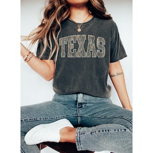 K&ouml;nnte beinhalten: Dunkelgraues T-Shirt mit dem Wort "TEXAS" in einem Camouflage-Muster auf der Brust. Das Shirt wird mit blauen Jeans und wei&szlig;en Turnschuhen kombiniert. Der Look wirkt l&auml;ssig und bequem, perfekt f&uuml;r den Alltag.