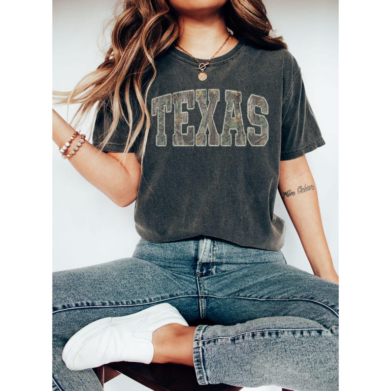 K&ouml;nnte beinhalten: Dunkelgraues T-Shirt mit dem Wort "TEXAS" in einem Camouflage-Muster auf der Brust. Das Shirt wird mit blauen Jeans und wei&szlig;en Turnschuhen kombiniert. Der Look wirkt l&auml;ssig und bequem, perfekt f&uuml;r den Alltag.