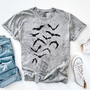 May include: A grey t-shirt featuring a black bat design, alongside light blue denim jeans and white high-top trainers. The shirt has a textured pattern. A casual, everyday outfit.
