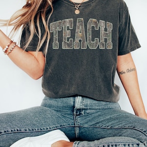 May include: Dark gray t-shirt with the word "TEACH" in a camouflage-style design. The shirt is paired with blue jeans and white slip-on shoes. The overall aesthetic is casual and comfortable.
