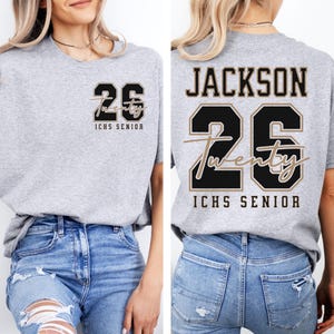 May include: A heather gray t-shirt with the number "26" in black and tan, and the word "Twenty" in cursive. The text "ICHS SENIOR" is below the number. The back of the shirt has the name "JACKSON" above the number.