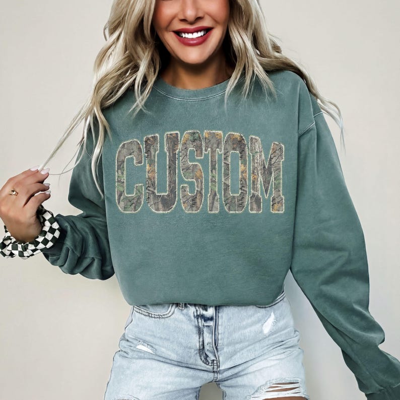 K&ouml;nnte beinhalten: Ein teal-farbenes Sweatshirt mit dem Wort "CUSTOM" in gro&szlig;en Blockbuchstaben. Die Buchstaben sind mit einem Camouflage-Muster gef&uuml;llt. Das Sweatshirt hat lange &Auml;rmel und einen Rundhalsausschnitt. Das Model tr&auml;gt Jeansshorts.