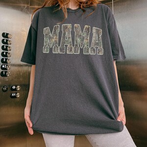 May include: Dark gray t-shirt with the word "MAMA" in a camouflage pattern across the front. The shirt has short sleeves and a relaxed fit. The text is in a bold, block letter style.