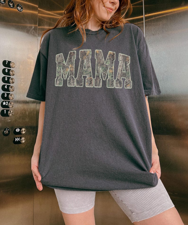 May include: Dark gray t-shirt with the word "MAMA" in a camouflage pattern across the front. The shirt has short sleeves and a relaxed fit. The text is in a bold, block letter style.