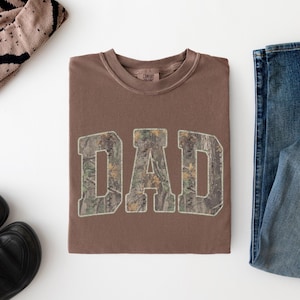 May include: A brown crewneck t-shirt featuring the word DAD in large block letters with a camouflage pattern fill. The shirt has a ribbed collar and is displayed alongside a pair of blue denim jeans, black shoes, and a tan patterned beanie.