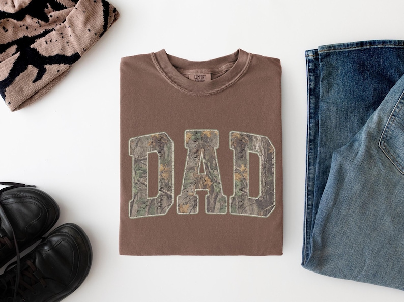 May include: A brown crewneck t-shirt featuring the word DAD in large block letters with a camouflage pattern fill. The shirt has a ribbed collar and is displayed alongside a pair of blue denim jeans, black shoes, and a tan patterned beanie.