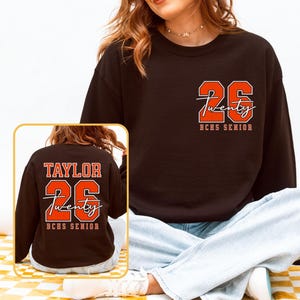 May include: Black sweatshirt with the number "26" and the word "Twenty" in a script font, with "BCHS SENIOR" below. The back of the sweatshirt has the name "TAYLOR", the number "26", the word "Twenty", and "BCHS SENIOR".