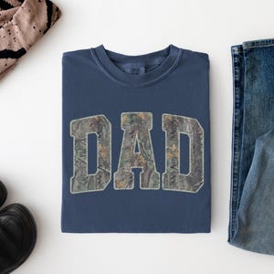 May include: A navy blue crewneck t-shirt featuring the word DAD printed in a camouflage pattern across the chest. The letters are in a bold, block font with a light-colored outline. The shirt is folded neatly and displayed on a plain white surface.
