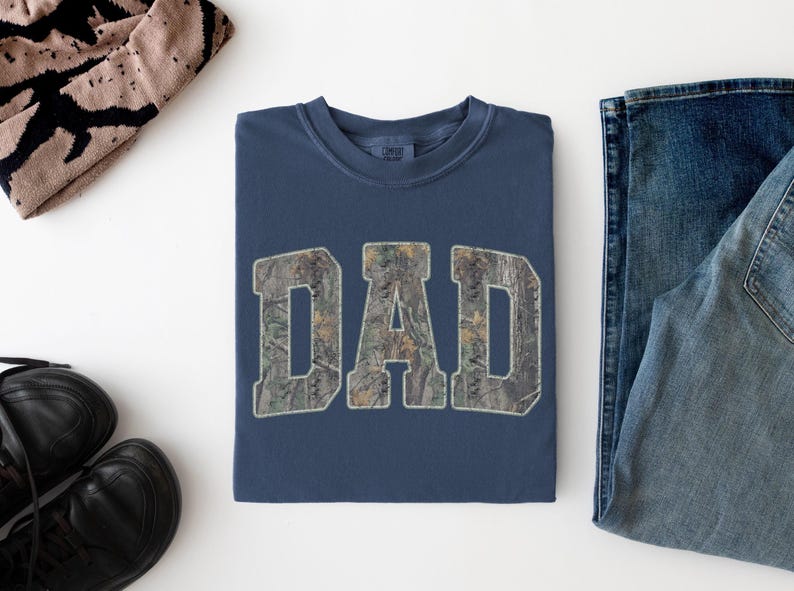 May include: A navy blue crewneck t-shirt featuring the word DAD printed in a camouflage pattern across the chest. The letters are in a bold, block font with a light-colored outline. The shirt is folded neatly and displayed on a plain white surface.