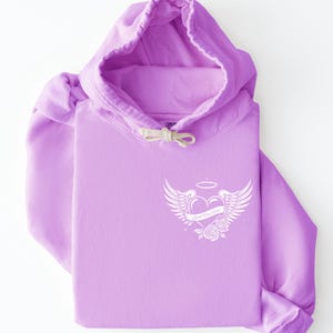 May include: A lavender hoodie with a white graphic on the chest. The graphic features a heart with wings, a halo, and roses, with the text "Good One Up". The hoodie has a drawstring hood and a kangaroo pocket.