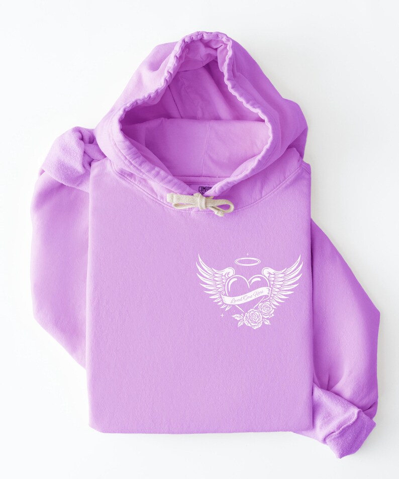May include: A lavender hoodie with a white graphic on the chest. The graphic features a heart with wings, a halo, and roses, with the text "Good One Up". The hoodie has a drawstring hood and a kangaroo pocket.