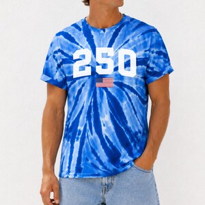 May include: A blue tie-dye t-shirt featuring the number "250" in white, with a small American flag graphic. The shirt is worn with denim shorts. The style is casual and suitable for summer.