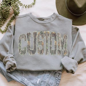 K&ouml;nnte beinhalten: Ein salbeigr&uuml;nes Sweatshirt mit dem Wort "CUSTOM" in gro&szlig;en Blockbuchstaben auf der Vorderseite. Die Buchstaben sind mit einem Camouflagemuster aus Gr&uuml;n-, Braun- und Beiget&ouml;nen gef&uuml;llt. Das Sweatshirt hat lange &Auml;rmel und einen Rundhalsausschnitt.
