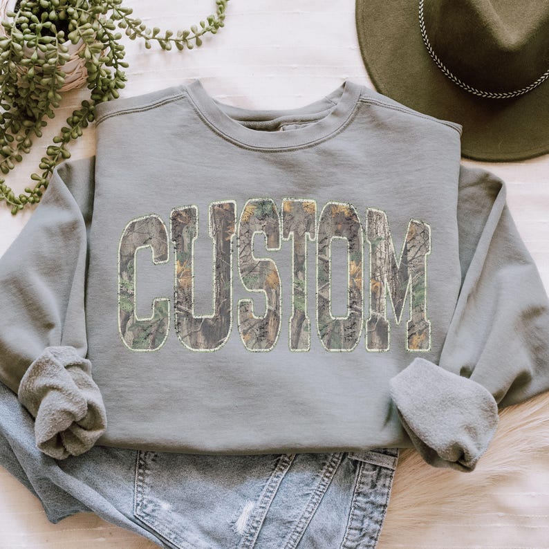 K&ouml;nnte beinhalten: Ein salbeigr&uuml;nes Sweatshirt mit dem Wort "CUSTOM" in gro&szlig;en Blockbuchstaben auf der Vorderseite. Die Buchstaben sind mit einem Camouflagemuster aus Gr&uuml;n-, Braun- und Beiget&ouml;nen gef&uuml;llt. Das Sweatshirt hat lange &Auml;rmel und einen Rundhalsausschnitt.