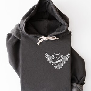 May include: Dark gray hoodie with a white drawstring and a white graphic on the chest. The graphic features a heart with wings, a halo, and the words "Loved One Here" on a banner, with roses below.