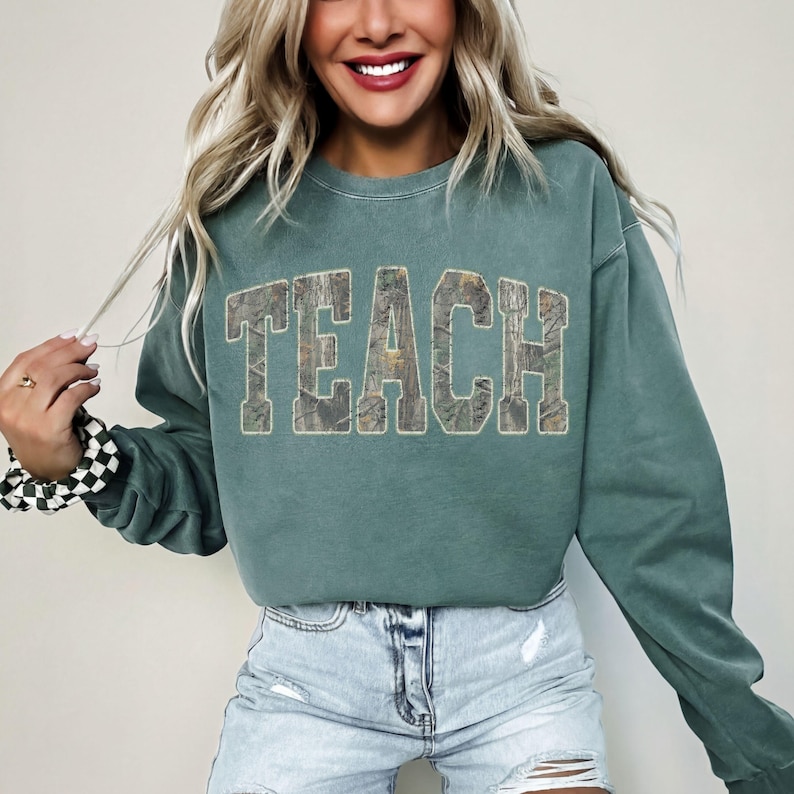 May include: Teal-colored sweatshirt with the word "TEACH" in a camouflage-style font. The sweatshirt has long sleeves and a crew neck. The model is wearing denim shorts and a black and white checkered scrunchie.