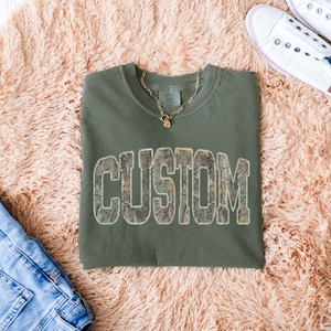 K&ouml;nnte beinhalten: Ein olivgr&uuml;nes Sweatshirt mit dem Wort "CUSTOM" in einem Tarnmuster auf der Vorderseite. Eine goldene Halskette und wei&szlig;e Turnschuhe sind ebenfalls zu sehen. Das Sweatshirt liegt auf einer rosa, flauschigen Oberfl&auml;che.
