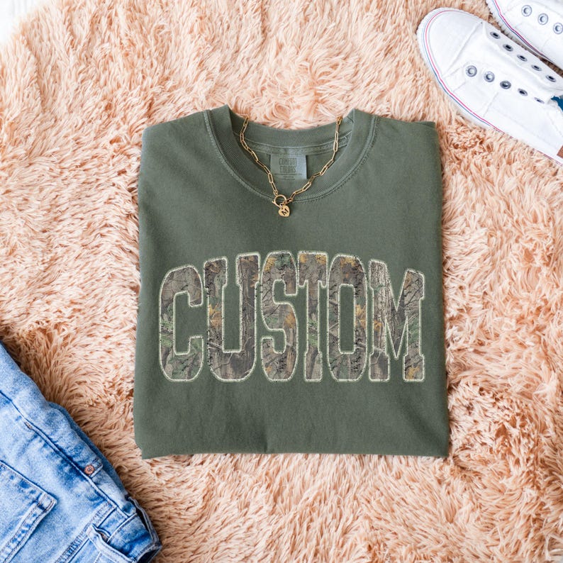 K&ouml;nnte beinhalten: Ein olivgr&uuml;nes Sweatshirt mit dem Wort "CUSTOM" in einem Tarnmuster auf der Vorderseite. Eine goldene Halskette und wei&szlig;e Turnschuhe sind ebenfalls zu sehen. Das Sweatshirt liegt auf einer rosa, flauschigen Oberfl&auml;che.