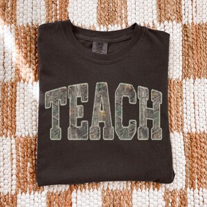May include: Dark brown t-shirt with the word "TEACH" in large, block letters. The letters have a camouflage pattern and a white outline. The shirt is made of a soft material and is likely a comfort fit.