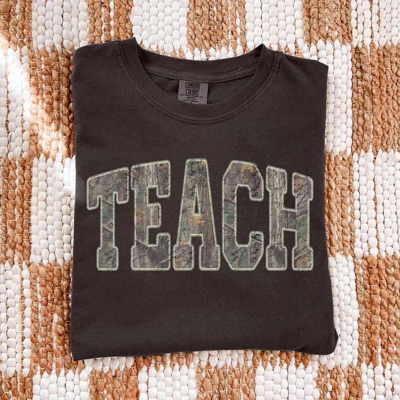 May include: Dark brown t-shirt with the word "TEACH" in large, block letters. The letters have a camouflage pattern and a white outline. The shirt is made of a soft material and is likely a comfort fit.