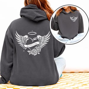 May include: Dark gray hoodie with a white graphic on the back featuring a heart, wings, roses, and the text "Loved One Here." A smaller version of the graphic is on the front. The hoodie has a drawstring hood.