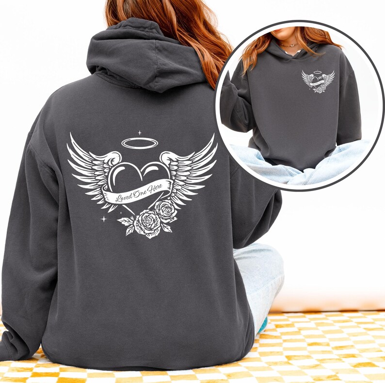 May include: Dark gray hoodie with a white graphic on the back featuring a heart, wings, roses, and the text "Loved One Here." A smaller version of the graphic is on the front. The hoodie has a drawstring hood.
