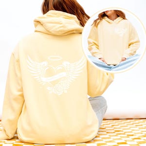 May include: A pale yellow hoodie with a white graphic on the back featuring a heart with wings, a banner, a halo, and roses. The front of the hoodie has a smaller version of the graphic. The hoodie has a hood and long sleeves.