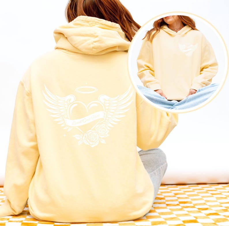 May include: A pale yellow hoodie with a white graphic on the back featuring a heart with wings, a banner, a halo, and roses. The front of the hoodie has a smaller version of the graphic. The hoodie has a hood and long sleeves.