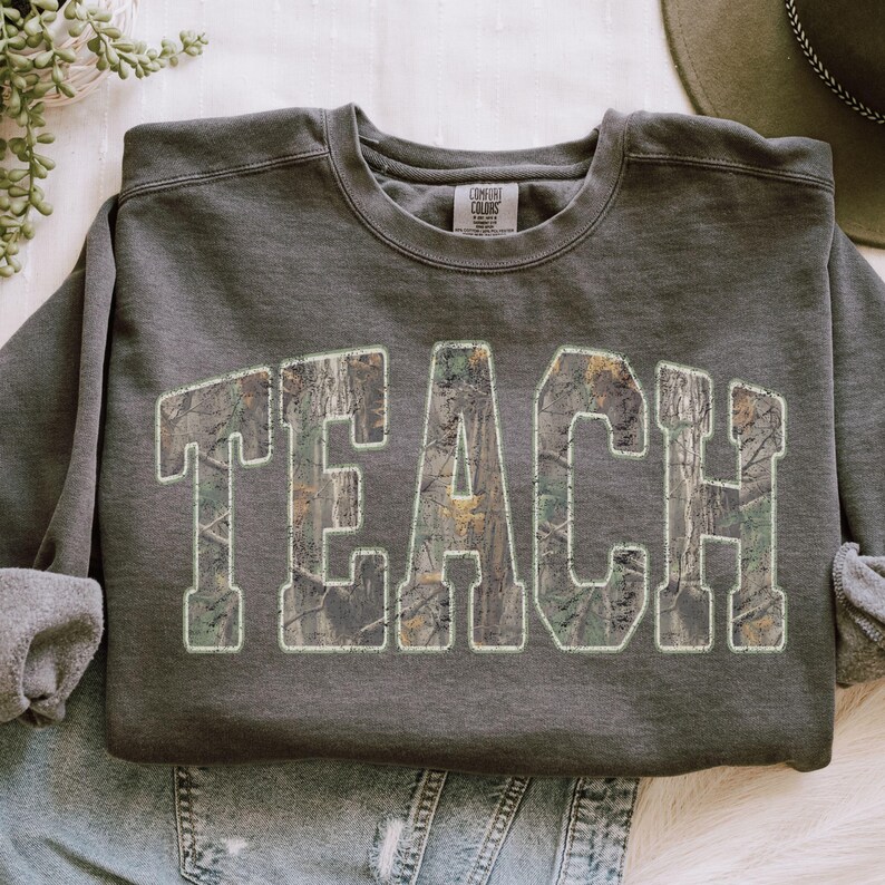 May include: Dark gray sweatshirt with the word "TEACH" in large, block letters. The letters are filled with a camouflage pattern and outlined in a light green color. The sweatshirt is folded, with the sleeves rolled up.