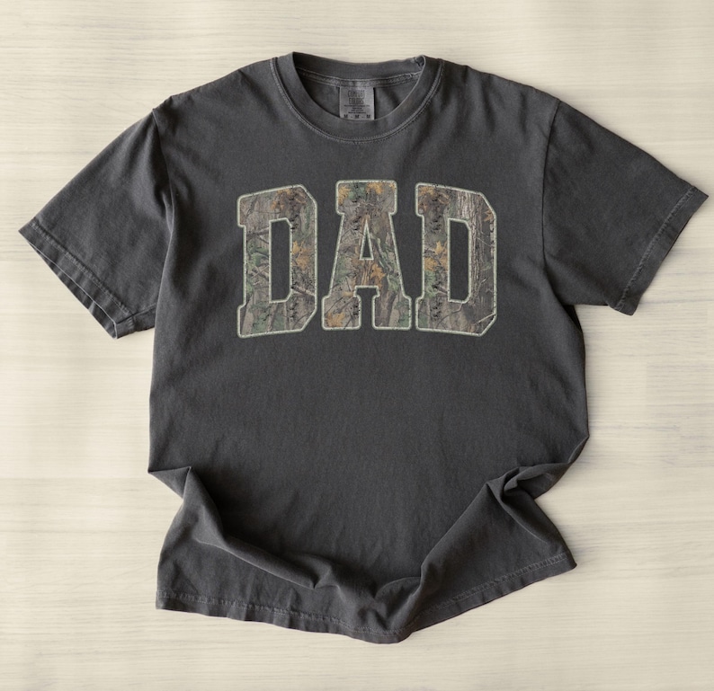 May include: A charcoal gray short-sleeve t-shirt featuring the word DAD printed in large, block-style letters across the chest. The letters are filled with a detailed forest camouflage pattern in shades of brown, green, and tan.